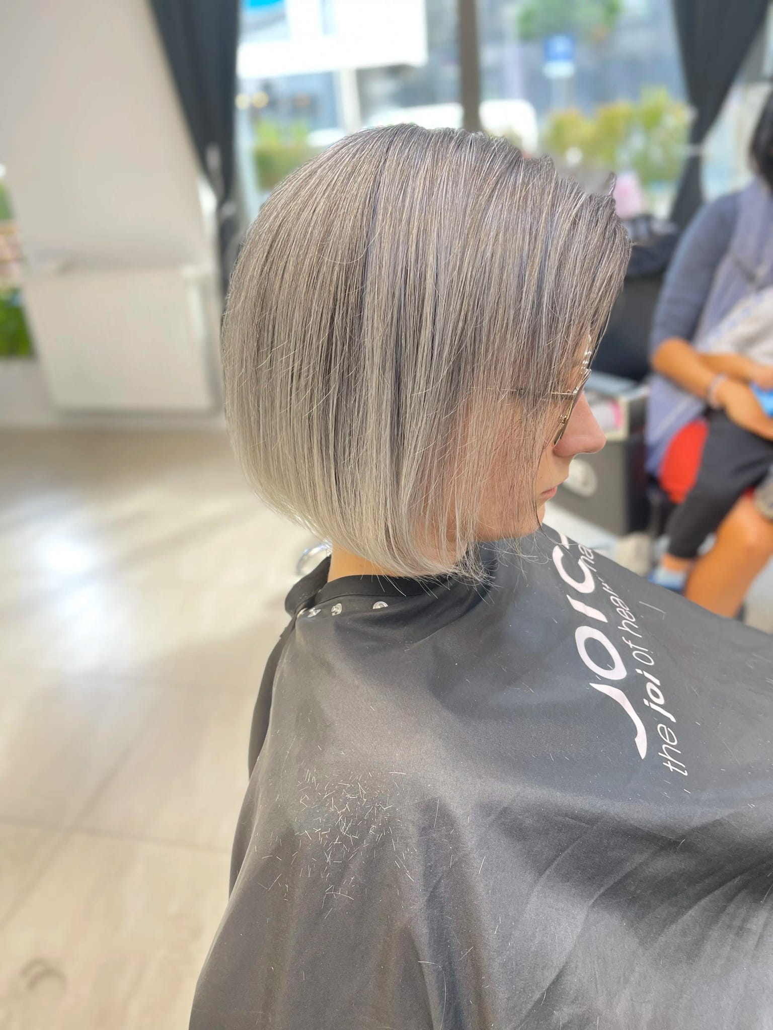 Osoba z gładką, srebrzystoszarą fryzurą typu bob sięgającą do brody, ubrana w czarną pelerynę fryzjerską z logo Joico, widoczna z profilu w salonie fryzjerskim.